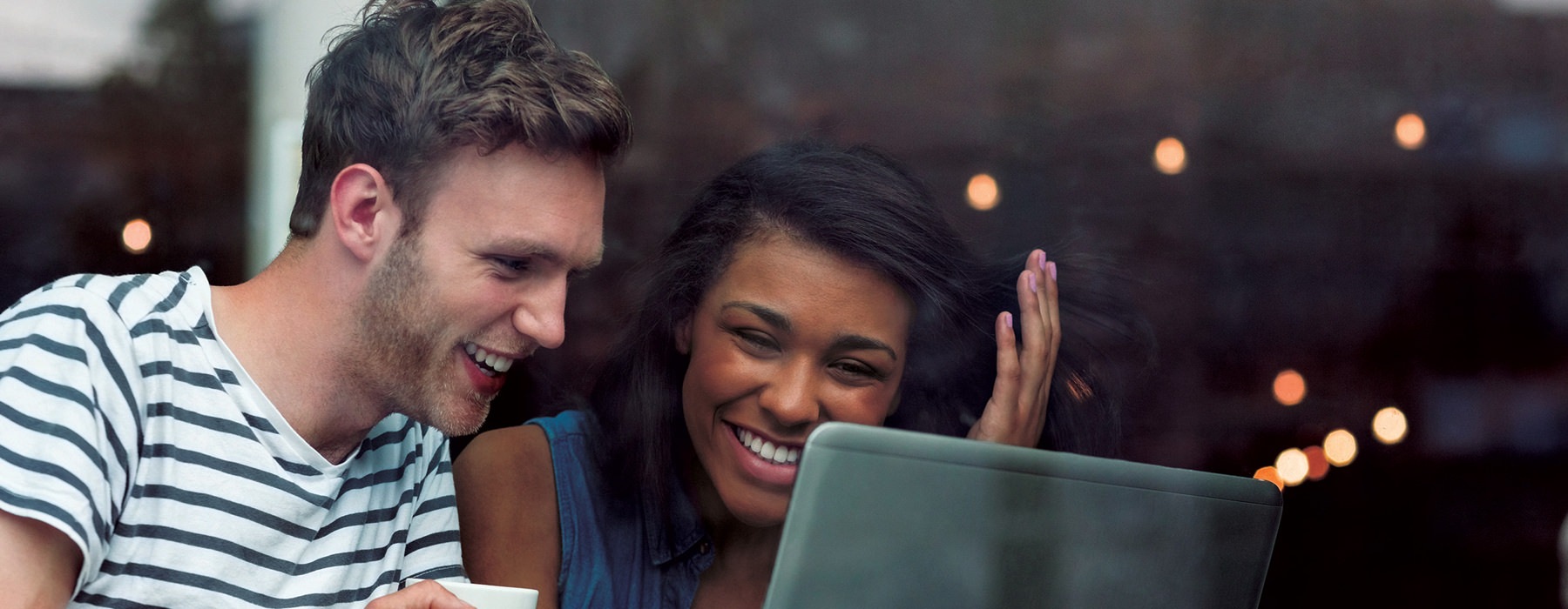 young man and woman in coffee shop window smile as they look at laptop screen